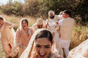 A joyous wedding party in blush pink and white outfits laughing together in a sunny outdoor field.