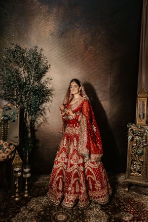 Elegant red and gold wedding lehenga photographed against a dark textured backdrop with decorative elements.