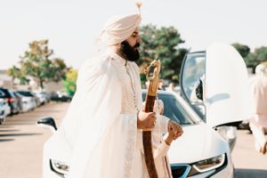 Person in traditional cream-colored attire stands next to a white luxury car in an outdoor parking lot.