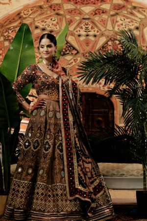 Traditional brown lehenga with intricate embroidery and sequin work shown against ornate wall with tropical plants.