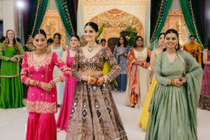 Wedding party members in vibrant Indian formal wear gather in an elegant ballroom with emerald drapes.