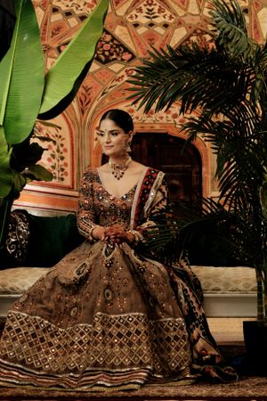 Traditional brown and gold bridal lehenga with intricate embroidery photographed against ornate architectural backdrop and tropical plants.