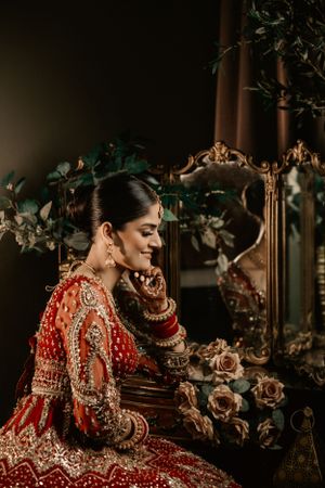 Traditional Indian bride in ornate red and gold lehenga poses elegantly beside an antique mirror with roses.