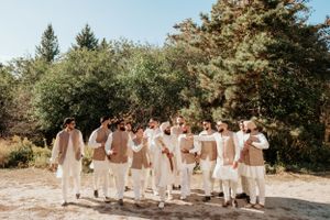 Wedding party in neutral beige and white attire stands together on sunny path surrounded by evergreen trees.