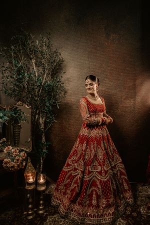 A traditional red and gold embroidered lehenga worn in an elegant indoor setting with lanterns and decorative foliage.