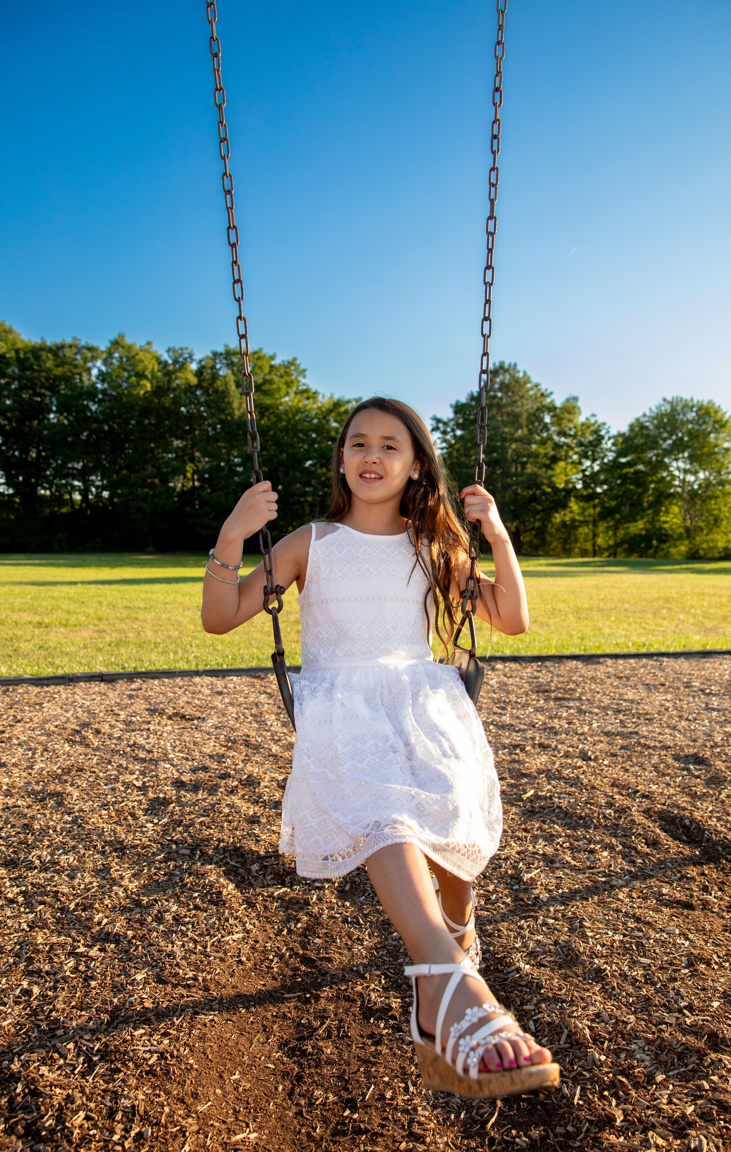 Someone wearing a white lace dress and sandals enjoys swinging at a playground on a sunny summer day.