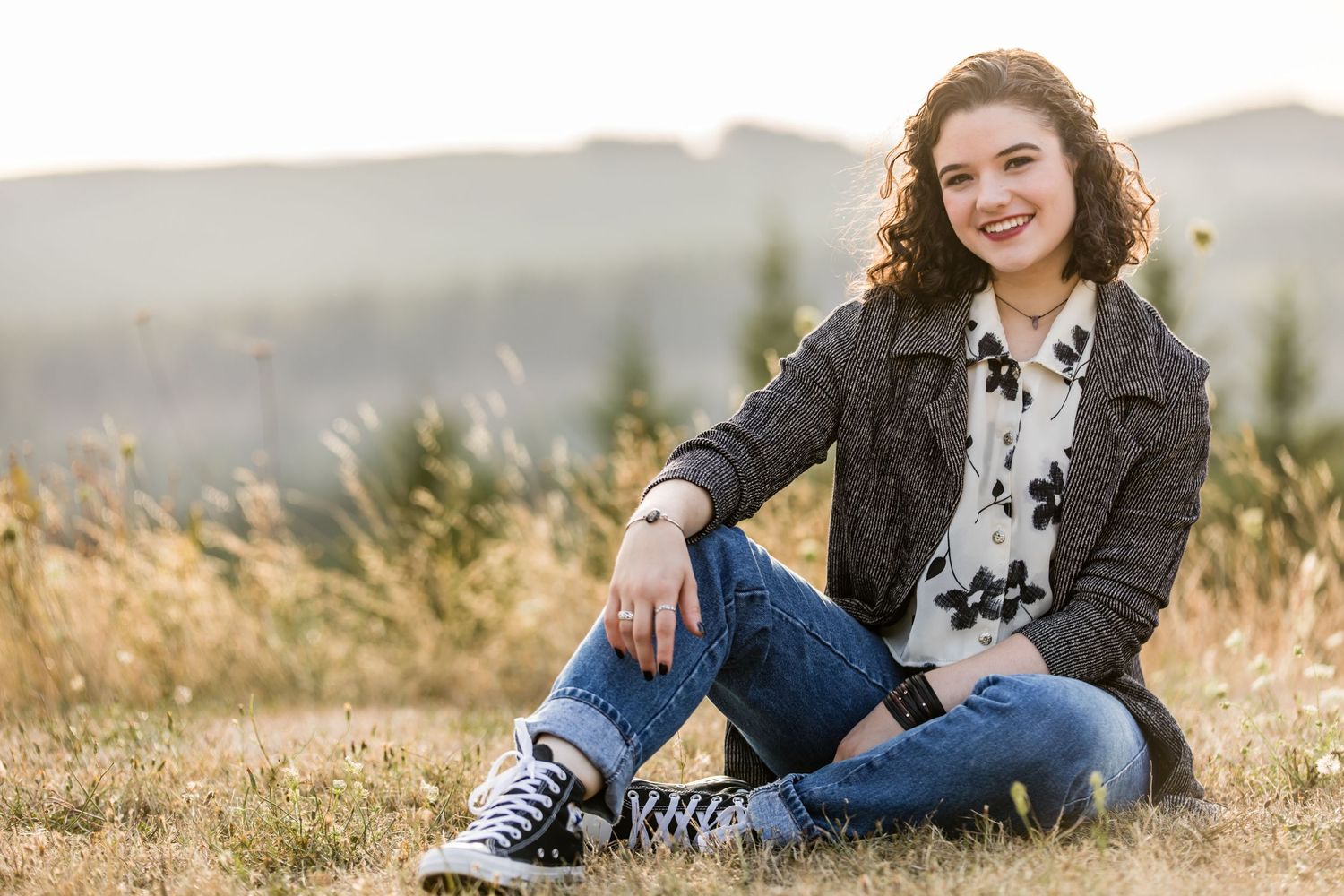 A person sits casually in a field wearing a dark jacket and jeans with mountains visible in the background.