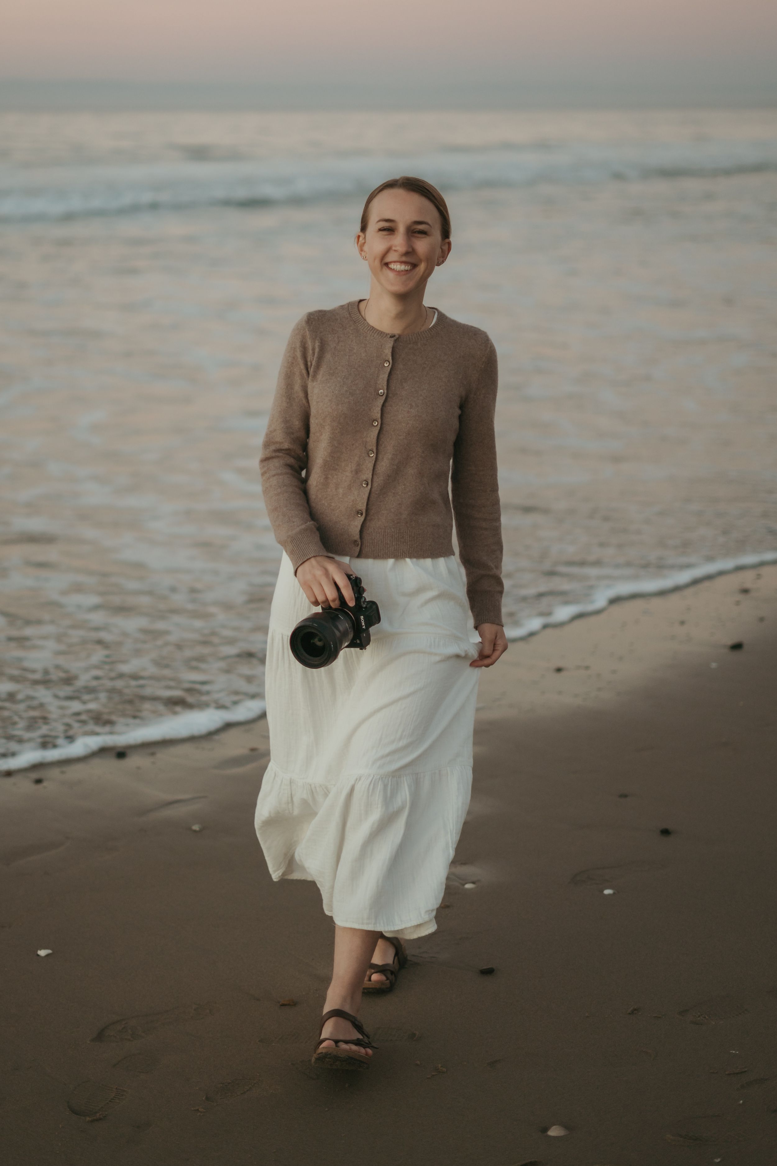 Someone in a white turtleneck sweater smiles while holding a camera outdoors near a wooden fence.