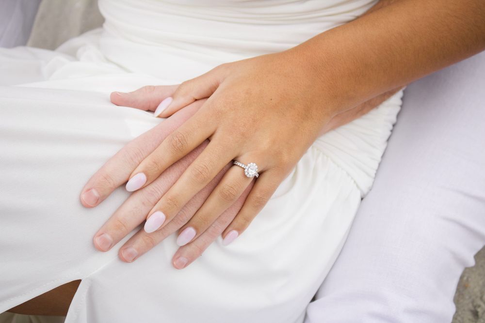 A manicured hand with a diamond engagement ring rests on white fabric showing off a classic bridal look.