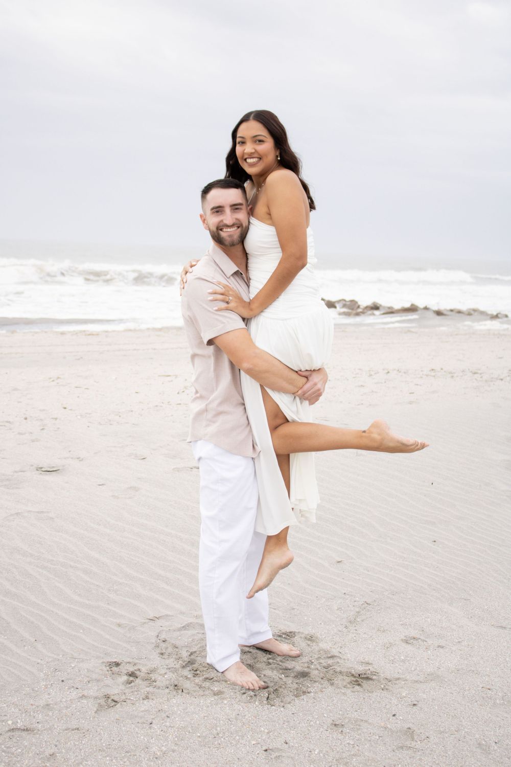 A couple in white outfits share a playful moment together on a cloudy beach.