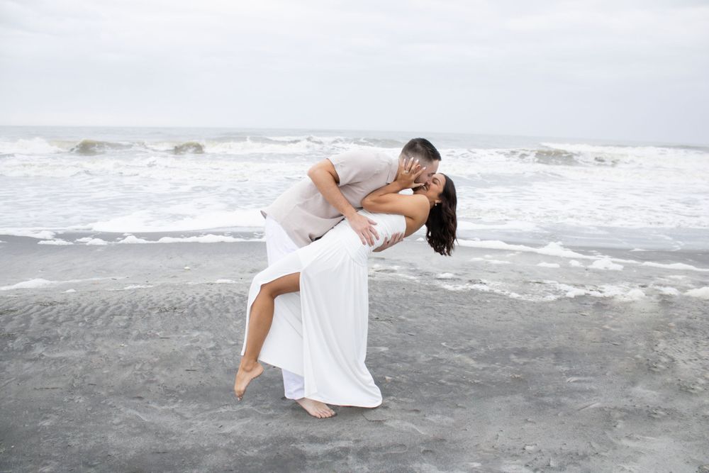 A romantic couple in white outfits share an intimate dance pose on a misty beach with crashing waves in the background.