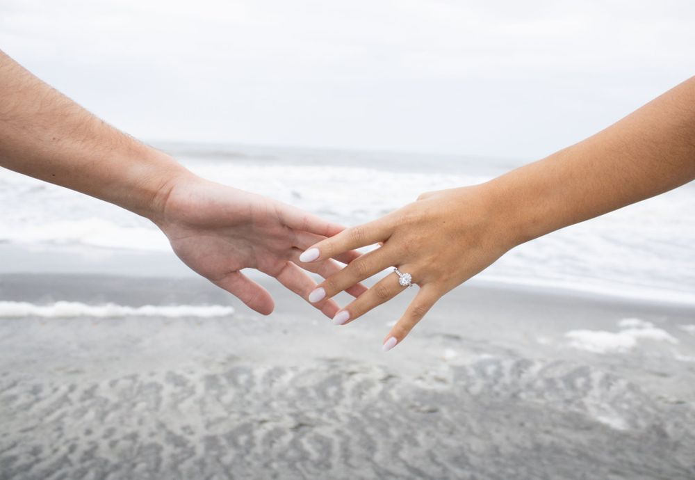 Two hands reaching out, fingertips touching tenderly against a blurred beach and ocean background.