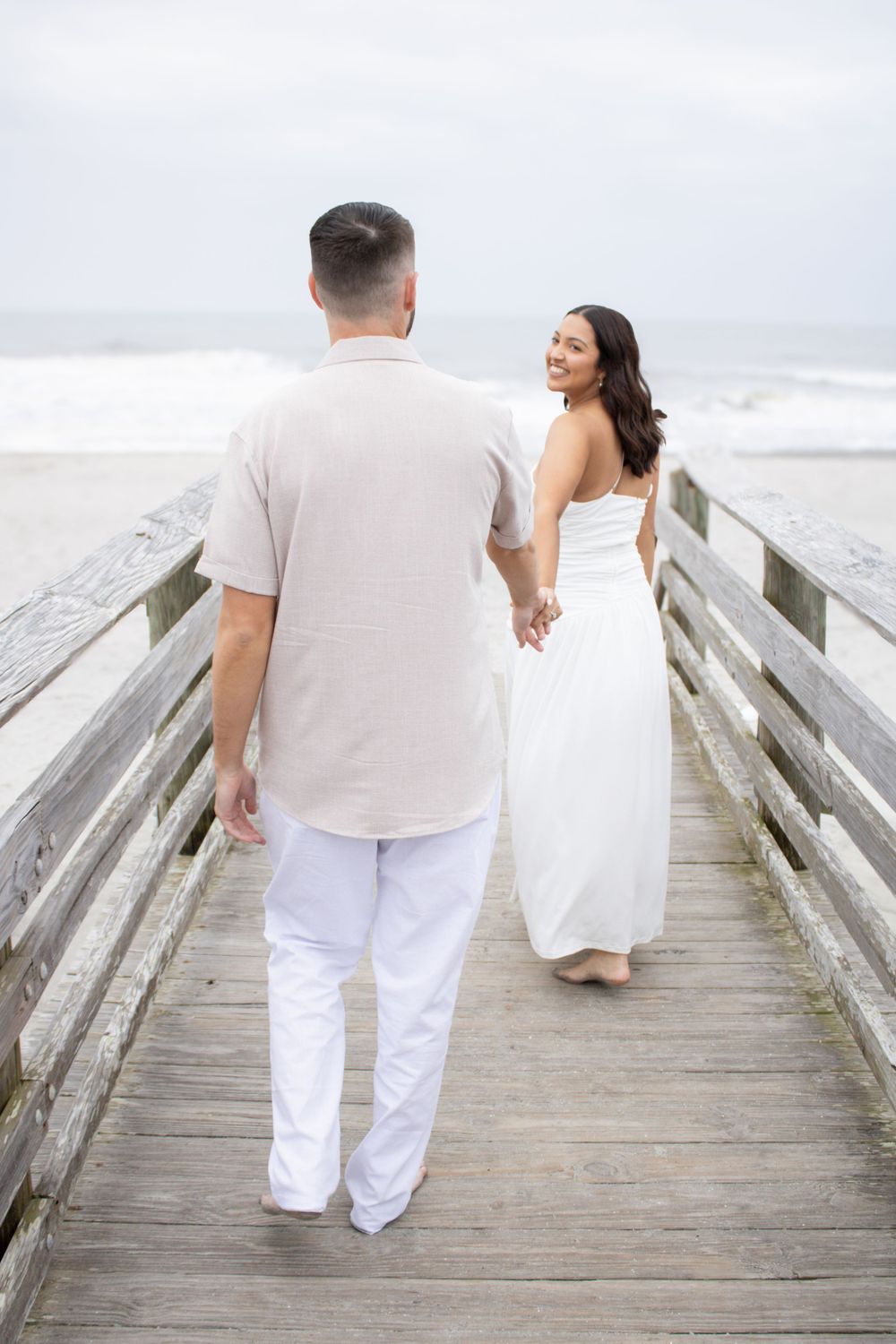 Couple in white outfits walking hand in hand on a wooden beach boardwalk with misty ocean backdrop.