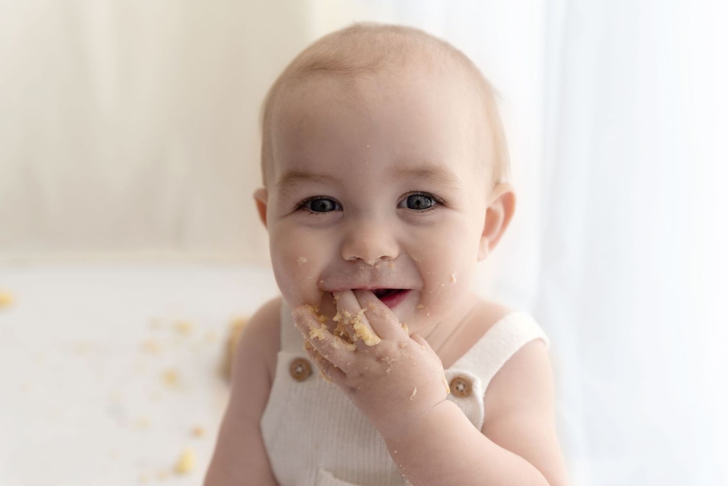 Baby in white clothing smiles while eating and making a happy mess during mealtime.