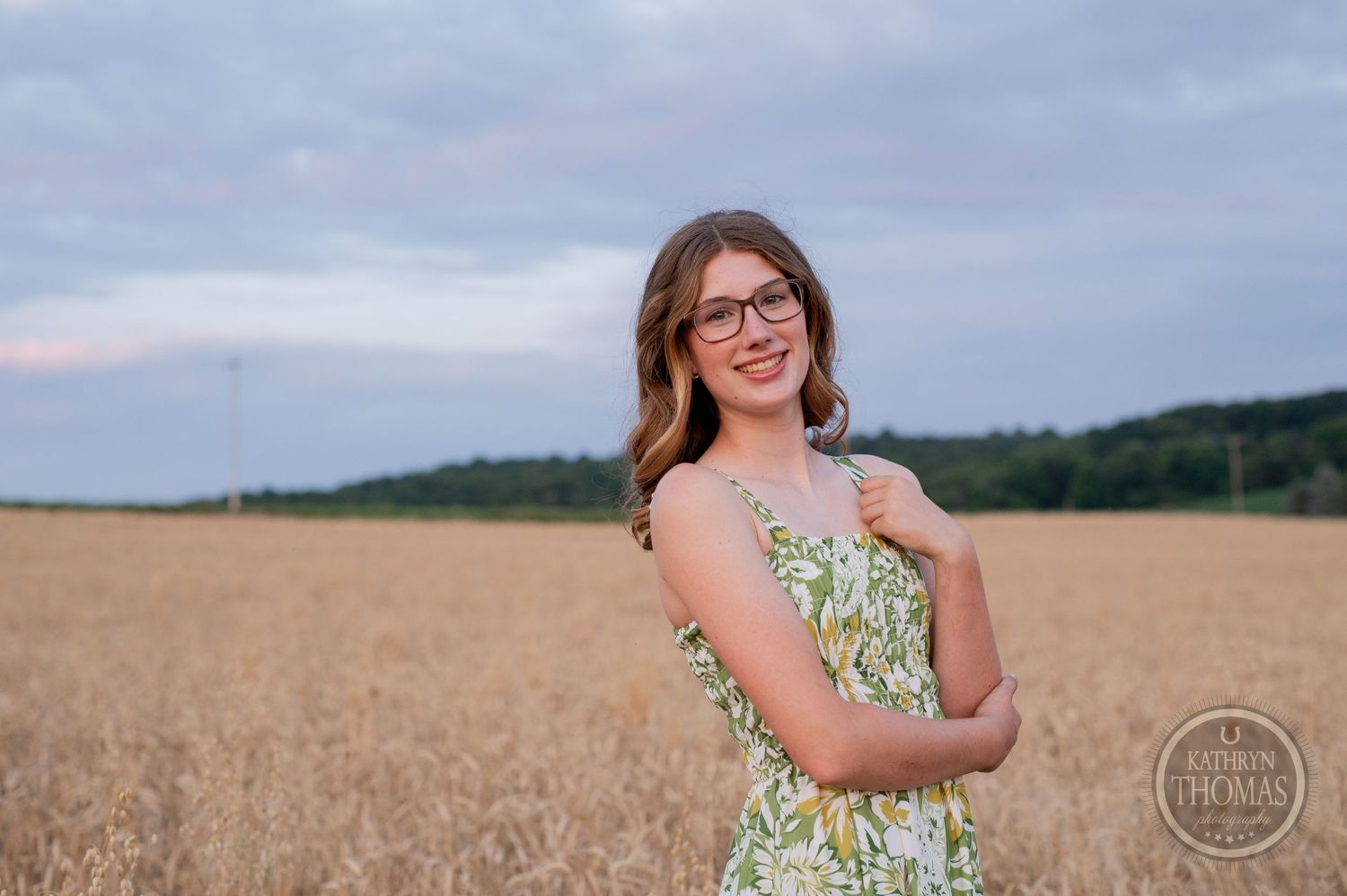 Person in floral dress and glasses poses cheerfully against evening sky in wheat field.