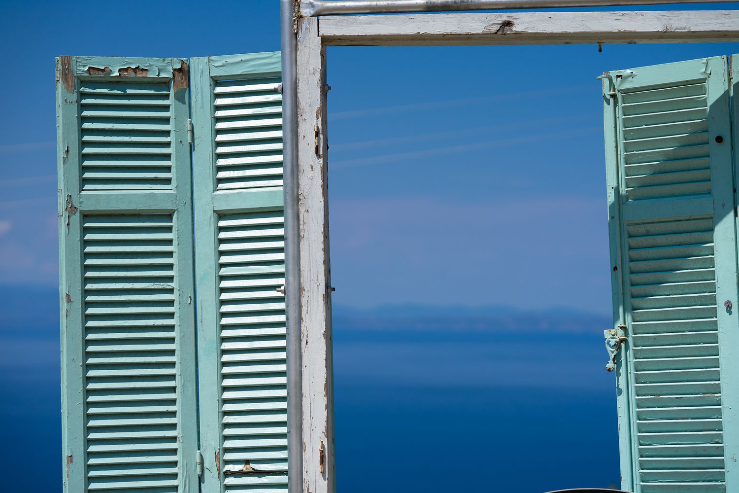Open mint green window shutters frame a stunning view of bright blue Mediterranean sea waters against a clear sky.