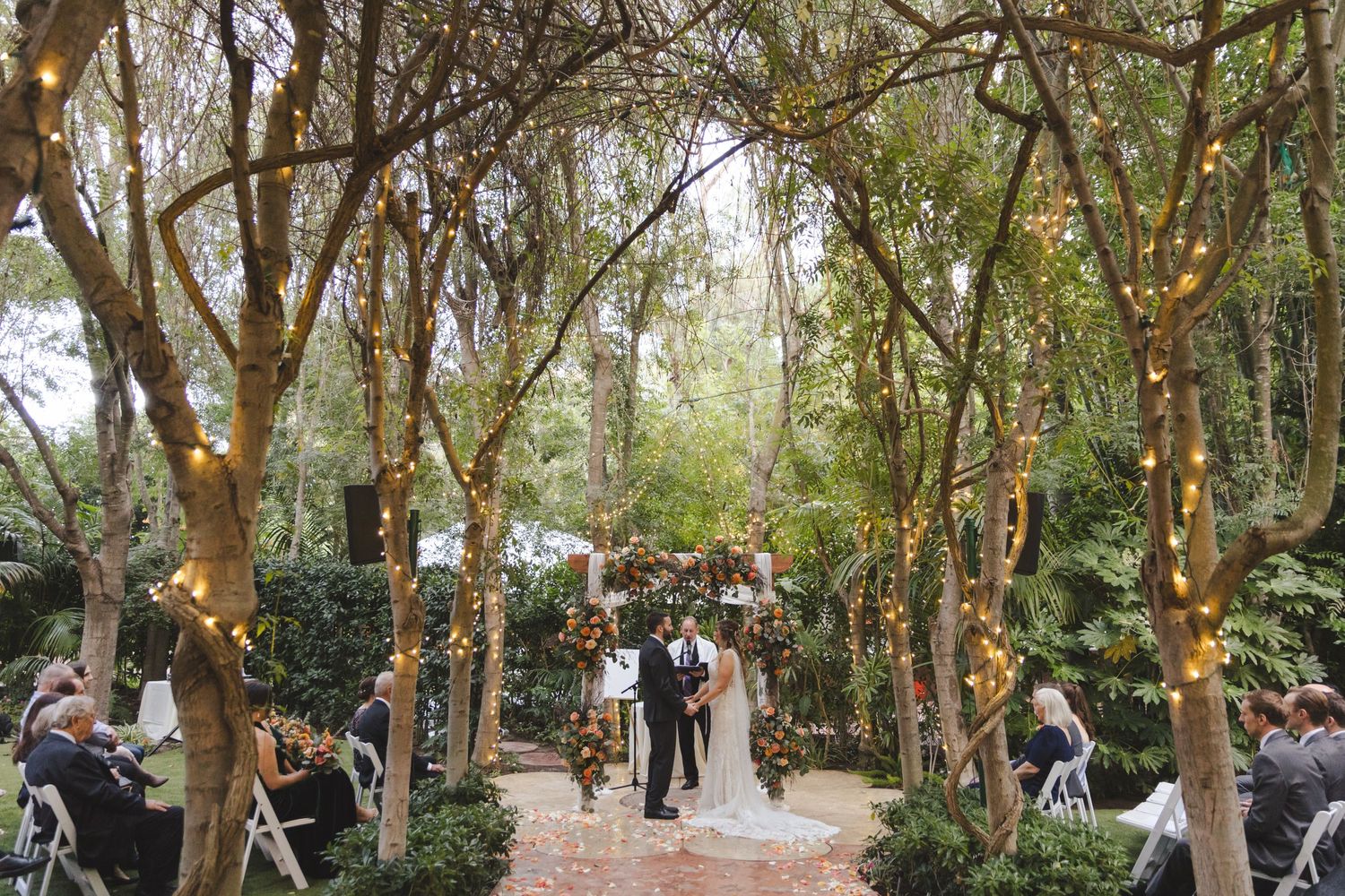 A romantic outdoor wedding ceremony takes place under a canopy of trees decorated with twinkling string lights.