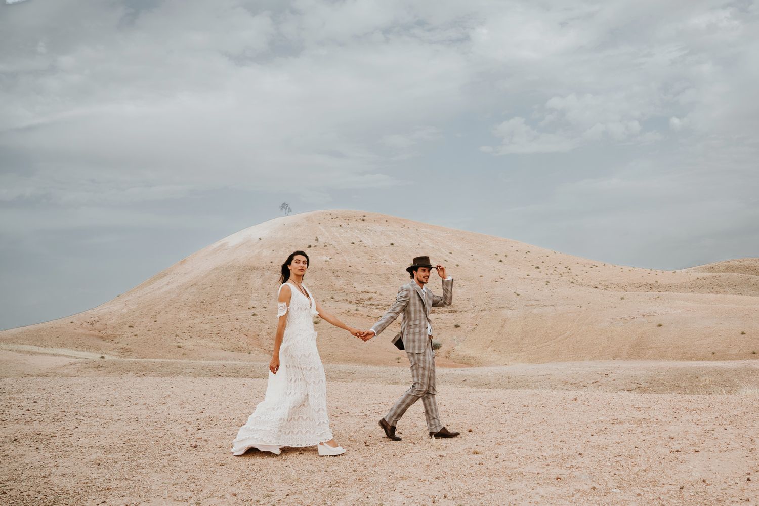 Two people in wedding attire walk hand in hand across a desert landscape with beige hills under cloudy skies.