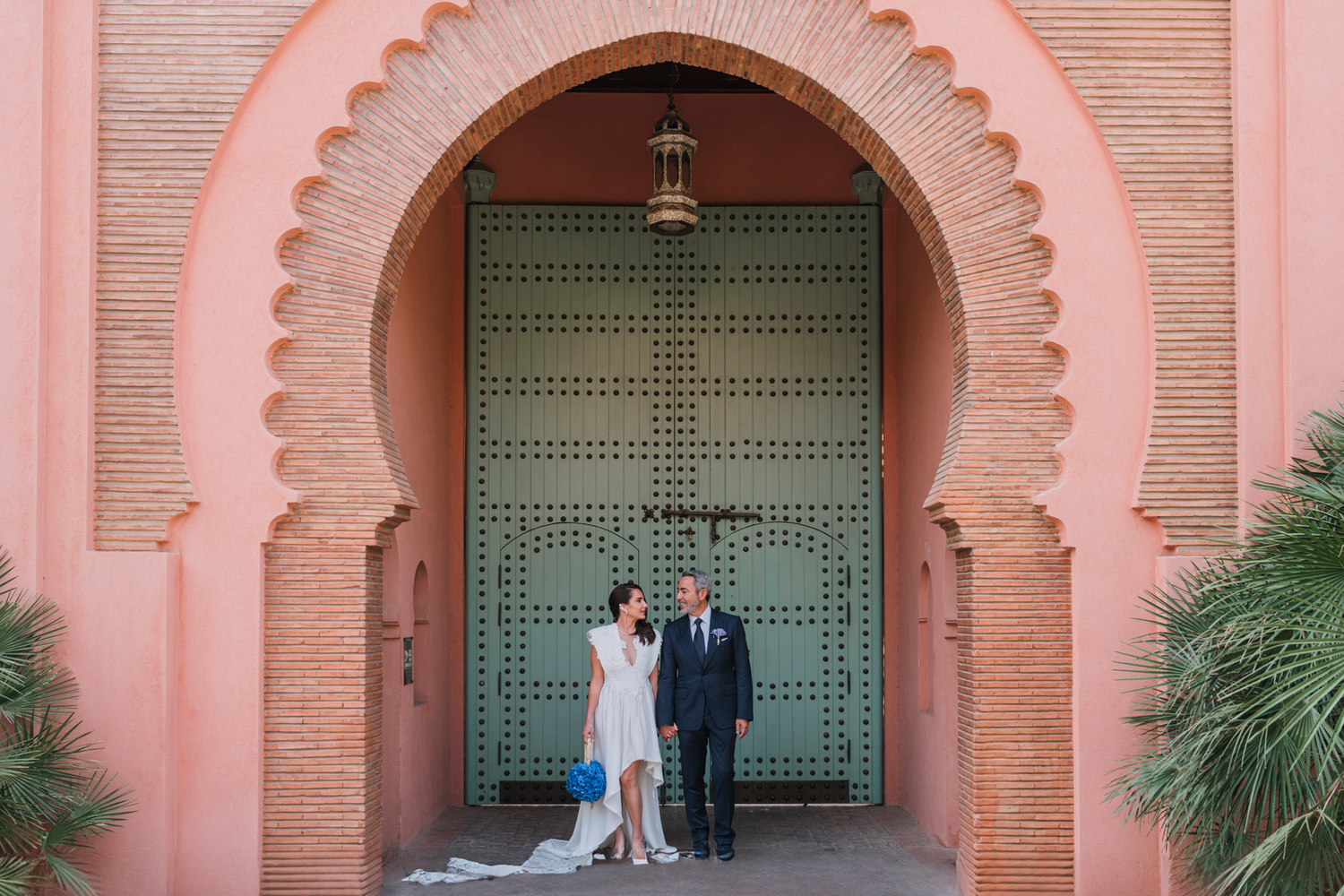 The couple stands under a Moroccan-style pink archway with ornate teal double doors behind them.