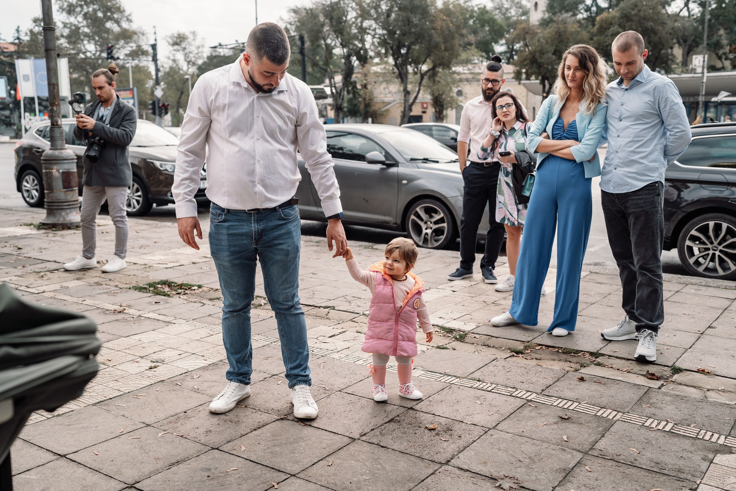 People gathered on a sidewalk outside with parked cars visible in the background.