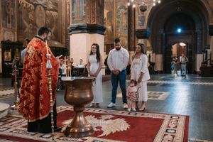 Priest in red robes conducting religious ceremony for family in ornate church interior.