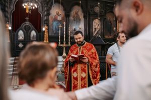 Inside an ornate Orthodox church, a baptism ceremony takes place with clergy in red robes and attendees in white.