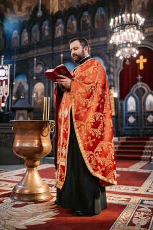 Orthodox priest in vibrant red and gold vestments performs ceremony on red carpet in ornate church interior.
