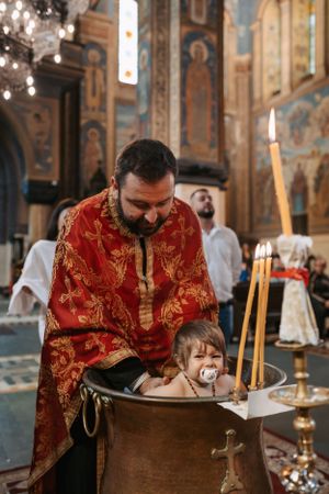 Orthodox priest in red vestments performs baptism ritual at a golden font in decorated church.