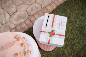 Gift box with strawberry decoration next to pink plate on grass.
