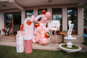 Party setup with pink balloon arch, strawberry decorations and white fountain.