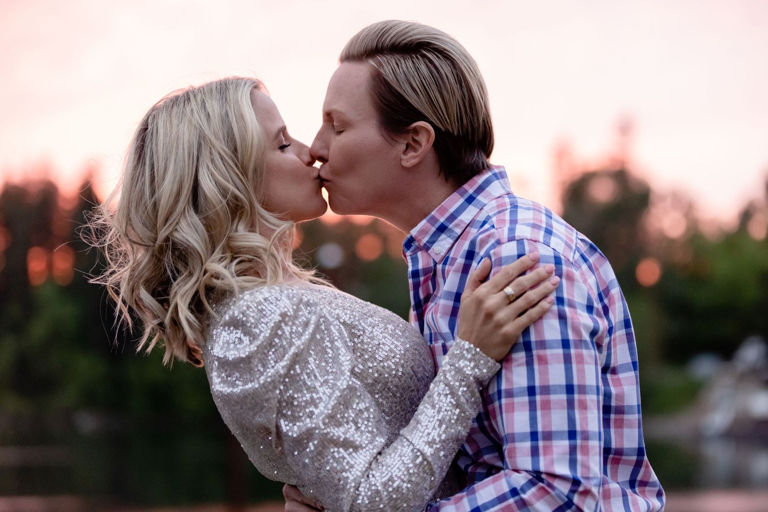 Couple sharing a sunset kiss by the water with warm lighting and trees in background.