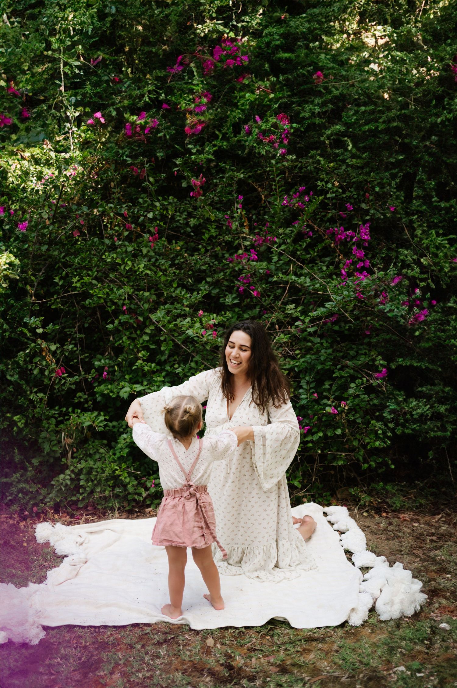 Two people sit together on a white blanket in front of blooming bougainvillea bushes with pink flowers in a garden setting.