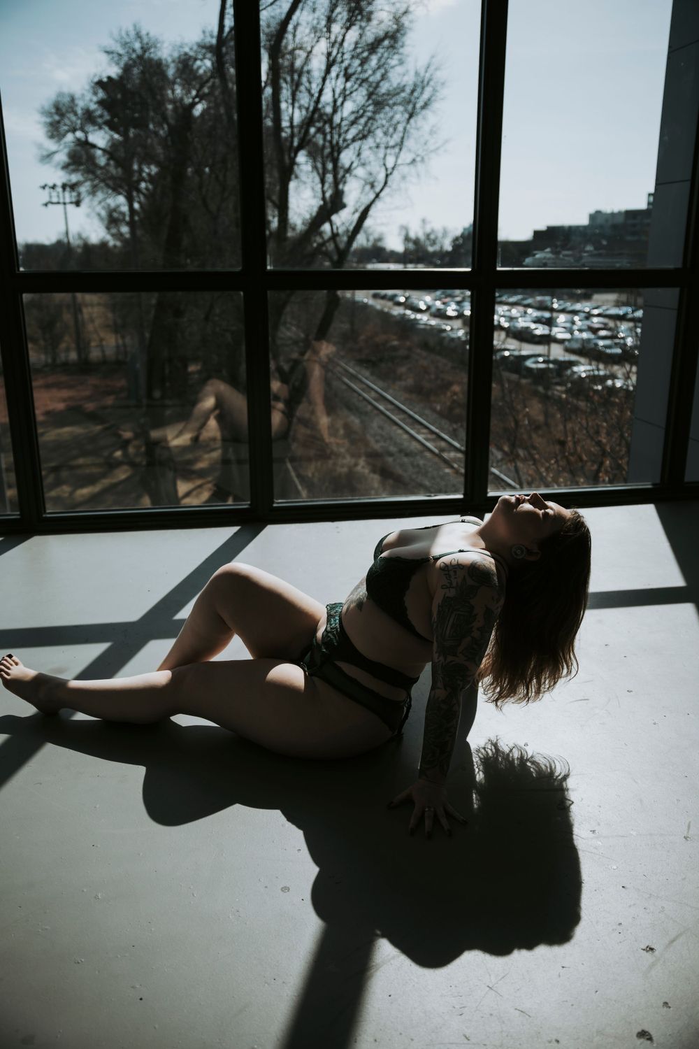 Silhouetted figure laying on sunlit floor by window overlooking train tracks and harbor marina in moody light.