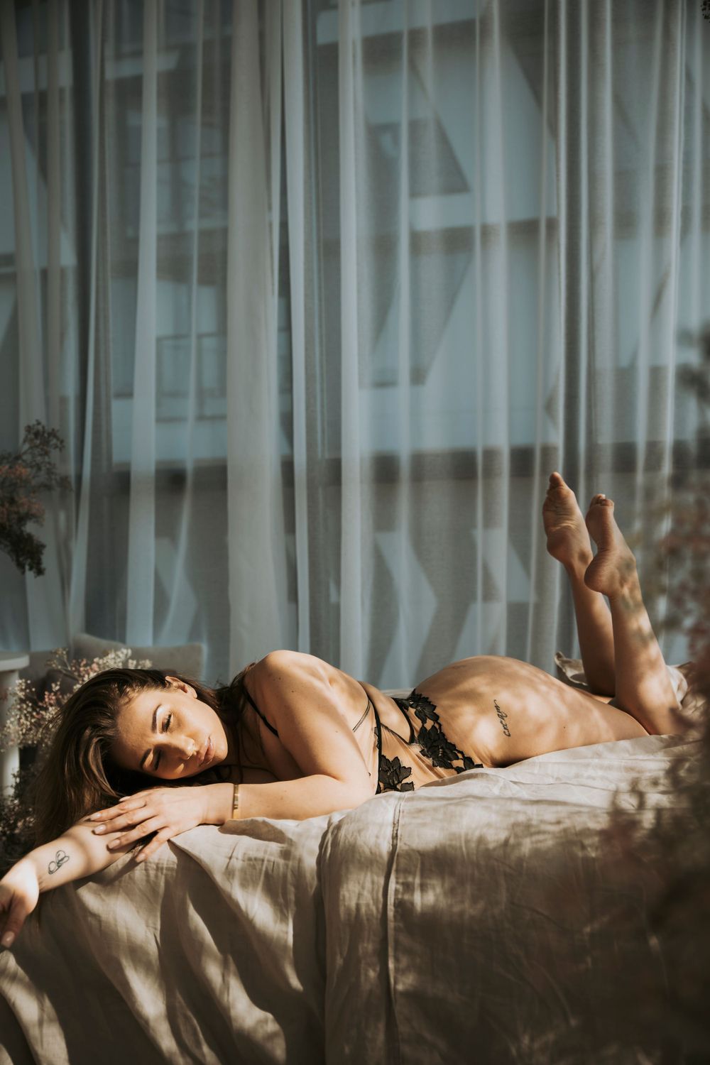 Artistic boudoir photo of a person resting on a bed in lingerie with soft natural window light streaming in.