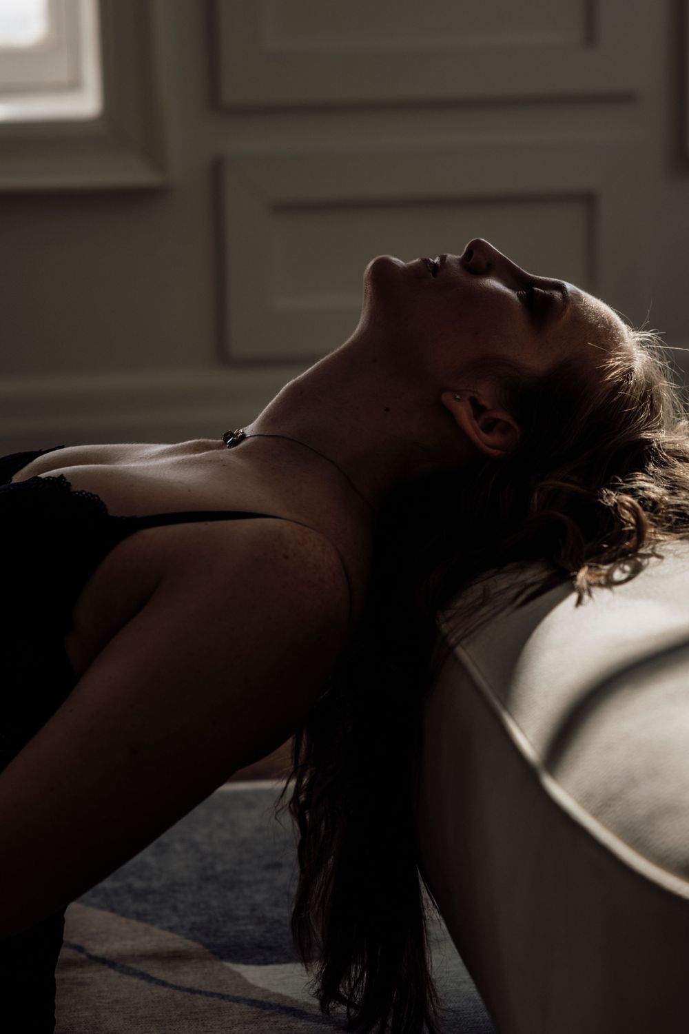 Figure silhouetted in dramatic lighting leans back on sofa, creating a moody artistic portrait.
