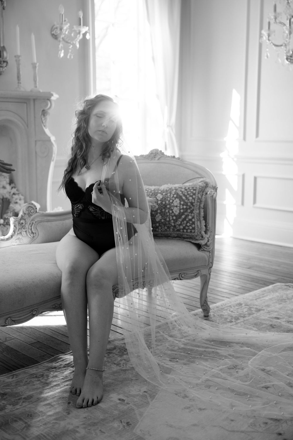 A moody black and white boudoir photo featuring flowing tulle fabric in an elegant room with chandeliers and antique furniture.