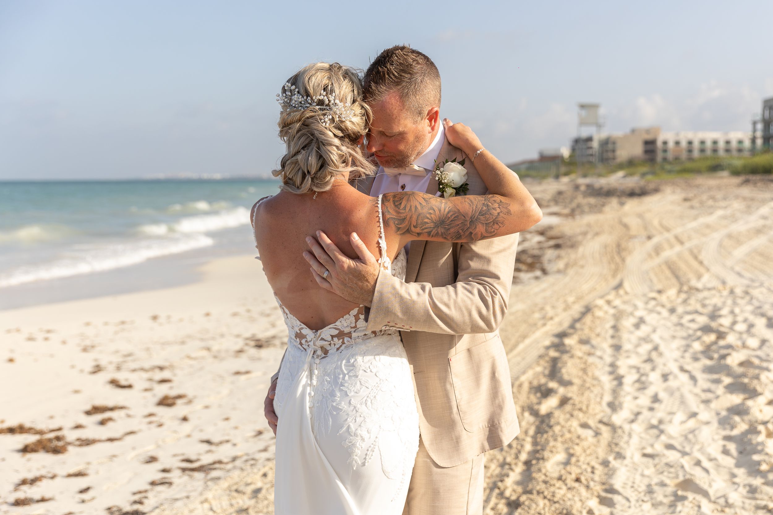 A romantic couple embraces on a sandy beach with the ocean and tall buildings visible in the background during a sunny day.