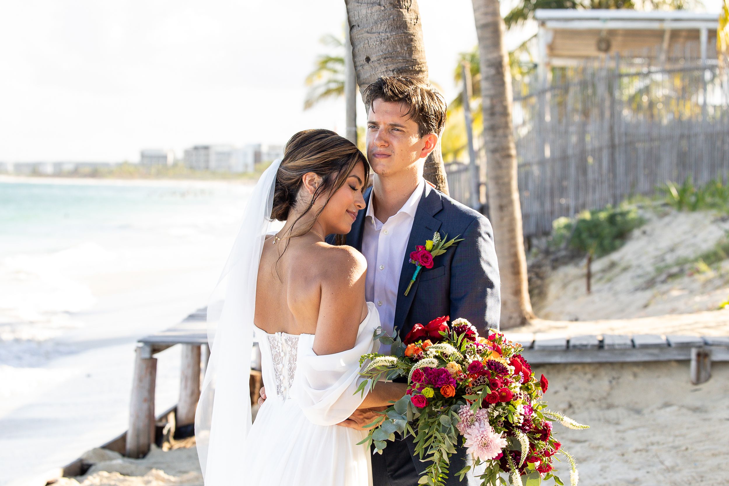 Bride and groom embrace on a tropical beach, bride holding vibrant red bouquet, wearing white gown with veil.