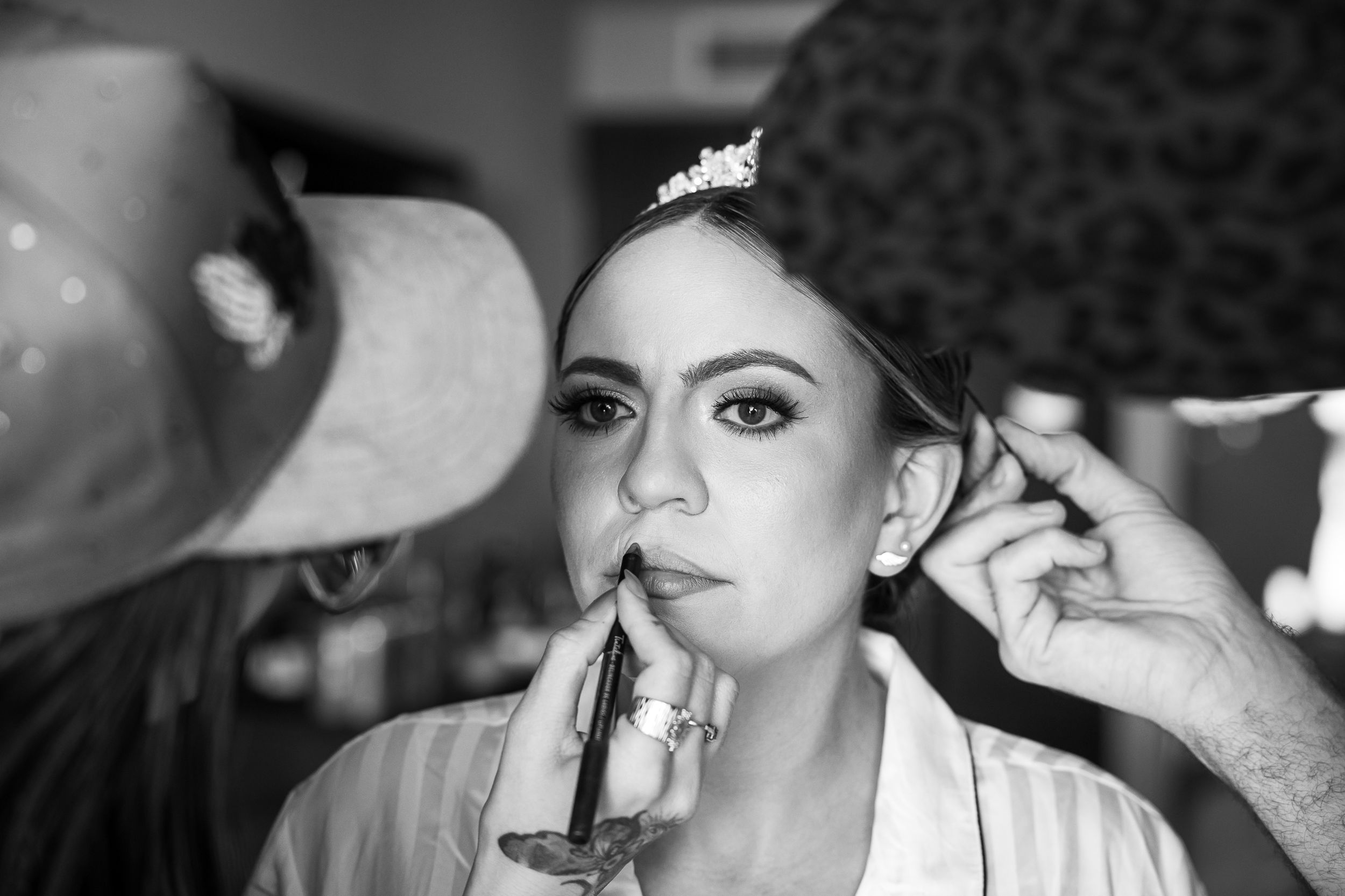 Black and white photo of a bride getting makeup applied, wearing a tiara and robe, with two makeup artists working on her face.