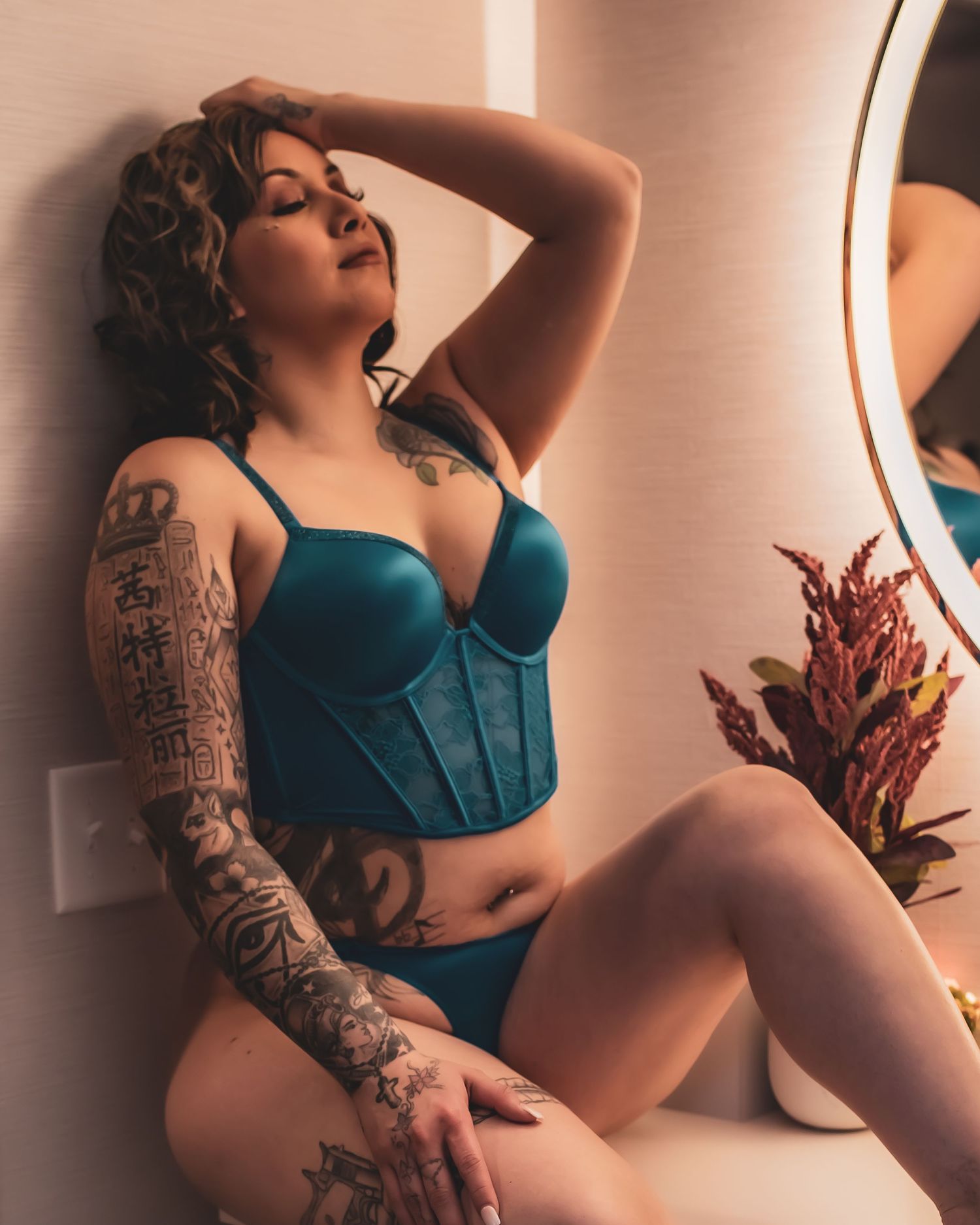 Figure in teal lingerie poses against light colored wall, with visible tattoo sleeve and dried flowers as props.