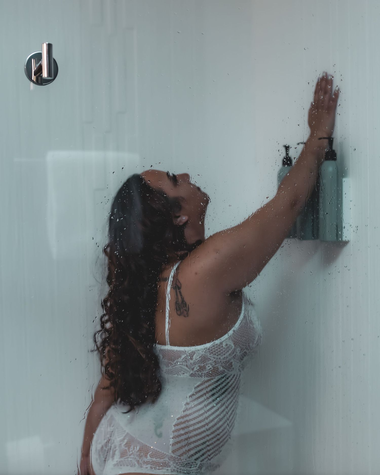 A figure in white lingerie stands against a foggy glass surface with arms raised upward.