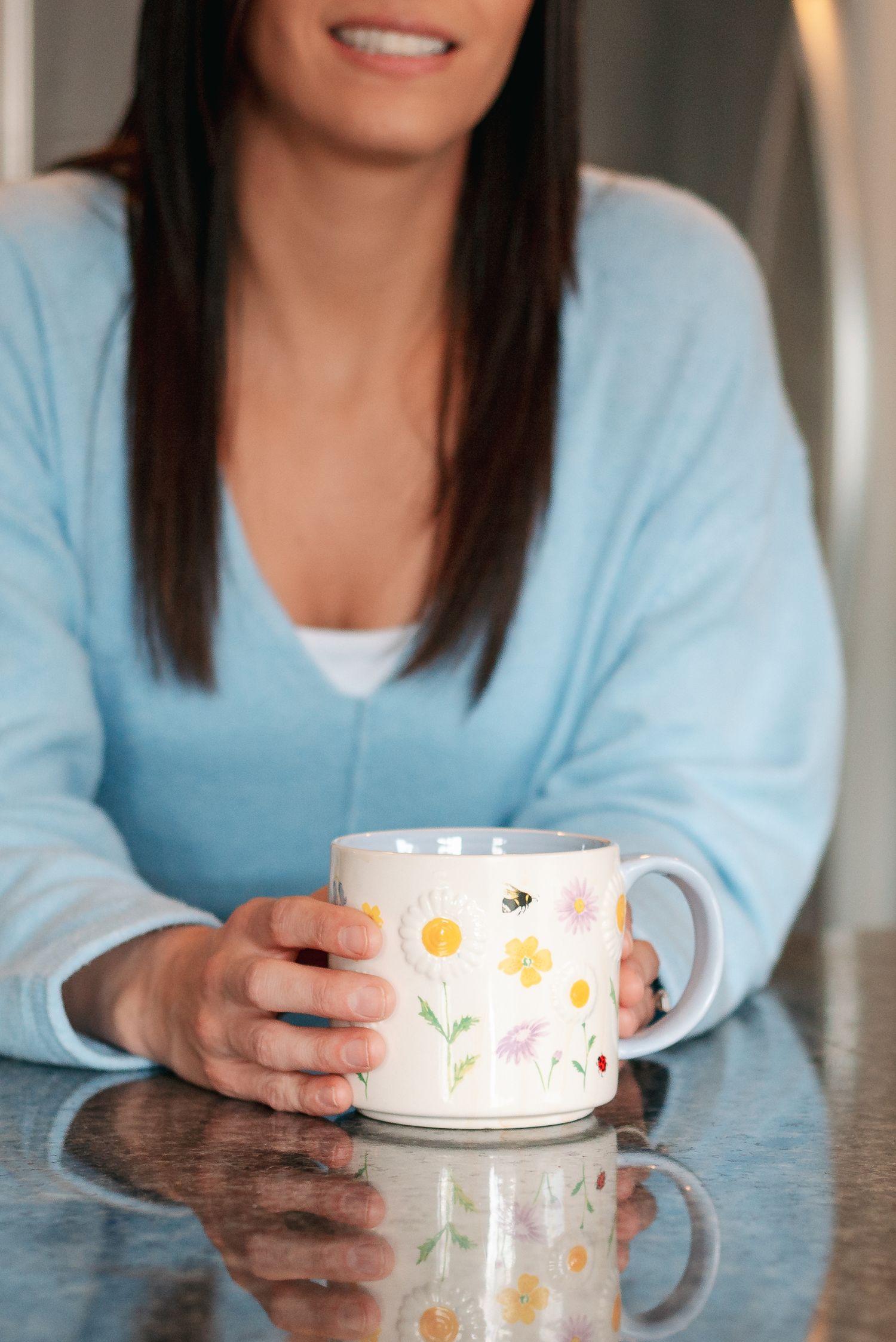 Someone in a light blue sweater holds a floral patterned coffee mug while sitting at a reflective table surface.