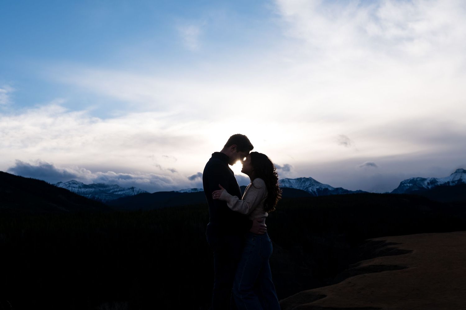 A scenic mountain landscape with evergreen trees silhouetted against misty peaks and cloudy skies in the distance.
