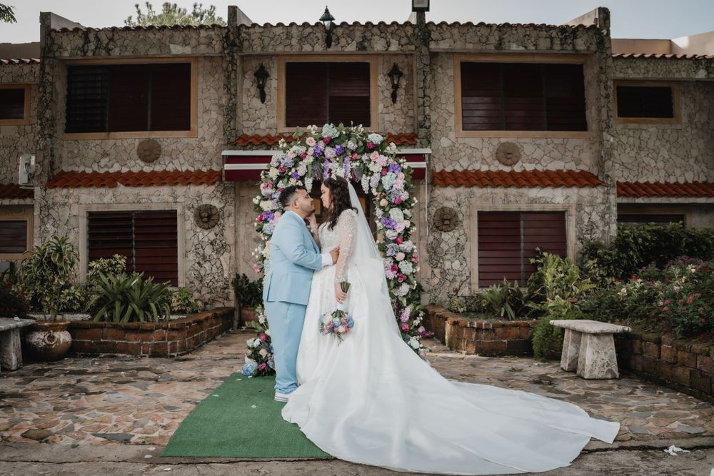 A wedding couple stands beneath a floral arch in front of a rustic stone building on a green carpet runner.