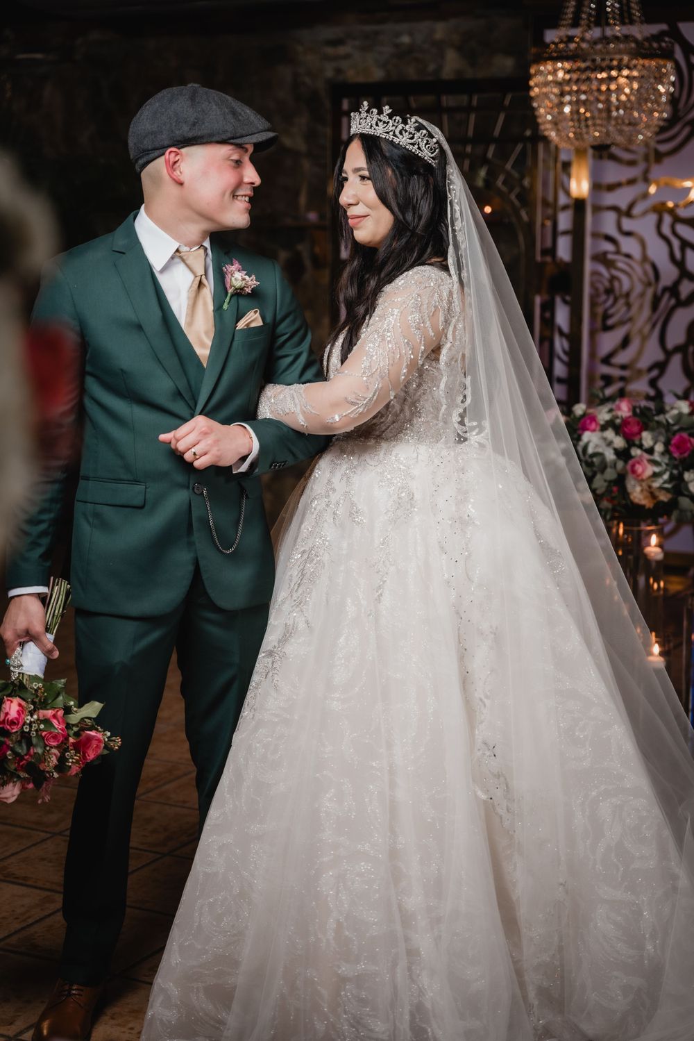 A couple shares a romantic moment at their wedding, with a green suit and lace gown under elegant chandeliers.