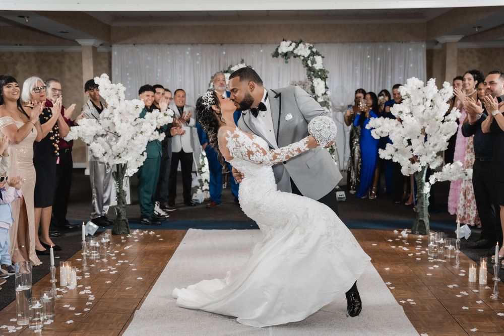 Newlyweds share a romantic dip on the dance floor surrounded by guests and white floral decorations at wedding reception.