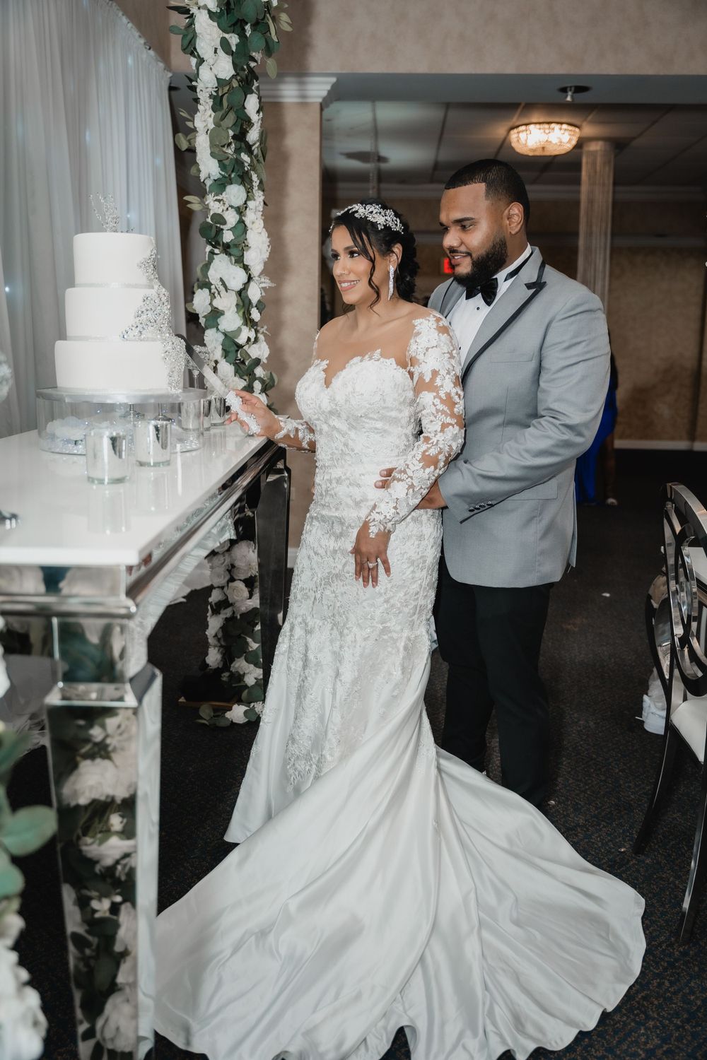A couple in elegant wedding attire stands next to a white tiered cake adorned with floral decorations.