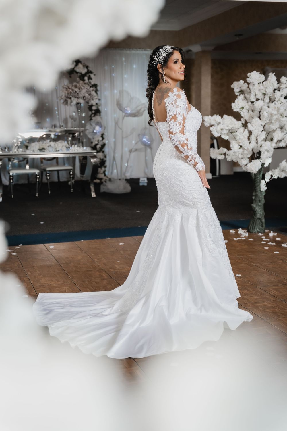A stunning white lace mermaid wedding gown with long sleeves and a flowing train on display in an elegant reception venue.