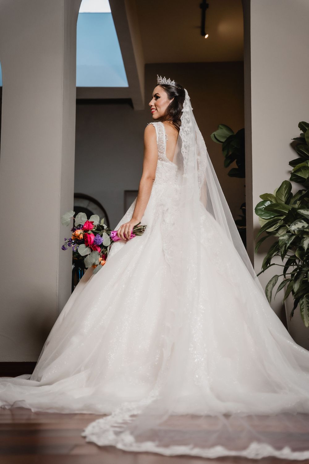 A bride in an elegant white ballgown wedding dress with long veil holds a colorful bouquet while standing in an elegant interior.