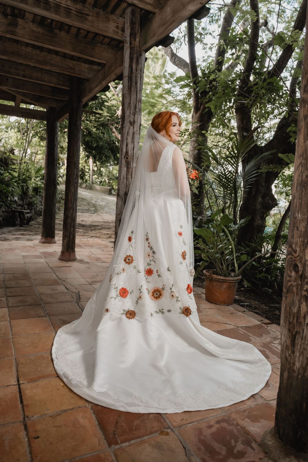 A wedding dress with embroidered floral details and long veil photographed on a terracotta patio with wooden beams overhead.